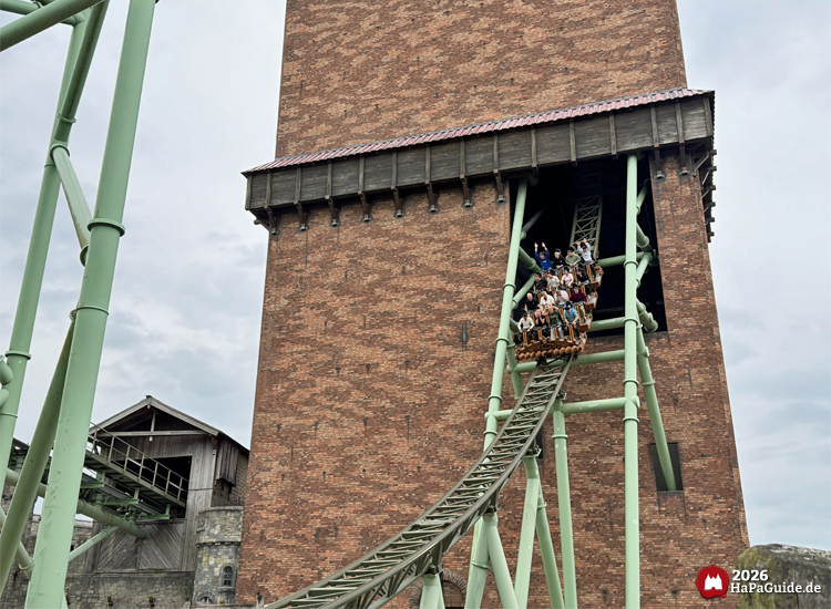 Zug der Achterbahn Der Schwur des Kärnan im Hansa-Park fährt aus dem hohen Backsteinturm heraus auf grünem Schienensystem, Fahrgäste heben die Arme während der Abfahrt