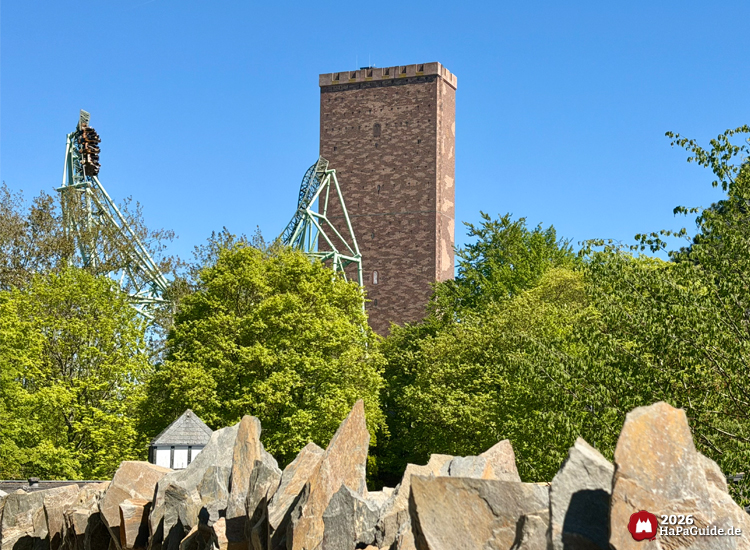 Backsteinturm der Achterbahn Der Schwur des Kärnan im Hansa-Park ragt über grüne Bäume und Felsen, im Hintergrund die grüne Stahlkonstruktion der Achterbahn unter blauem Himmel