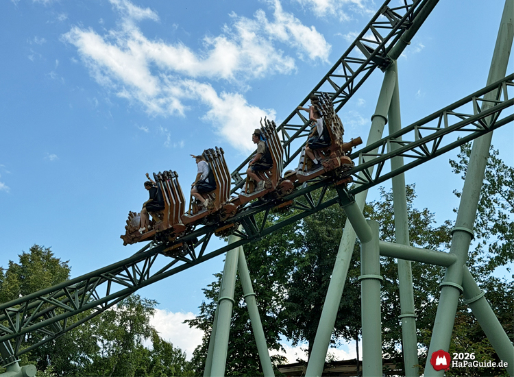 Zug der Achterbahn Der Schwur des Kärnan im Hansa-Park auf grünem Schienenabschnitt, Fahrgäste genießen die Fahrt unter blauem Himmel mit weißen Wolken