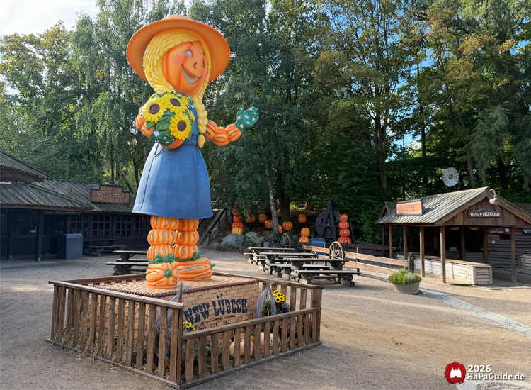 Große Kürbisfigur Martha Jane Field während Erntedank im Hansa-Park mit gelbem Hut und blauer Kleidung, hält Sonnenblumen und grünes Blatt, steht im Westernbereich New Lübeck zwischen Holzhütten und Bäumen