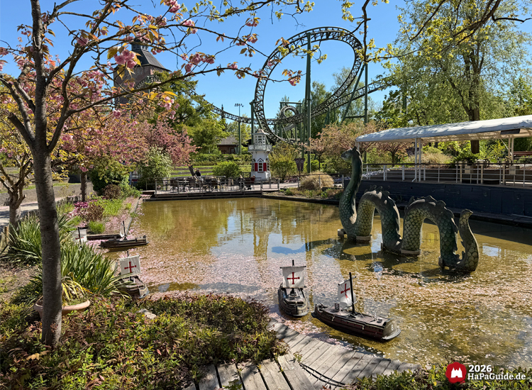 Teich mit Fernlenkschiffen und grüner Seeschlange Nessie im Hansa-Park, umgeben von blühenden Bäumen und der Achterbahn Flucht von Novgorod im Hintergrund