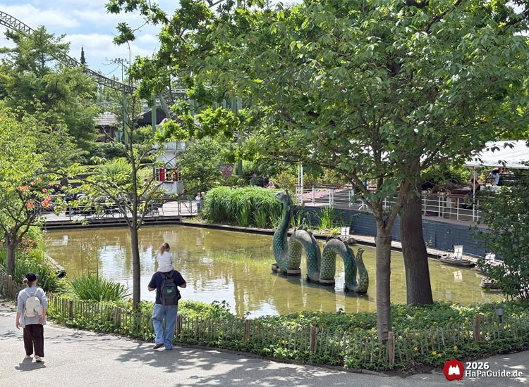 Teich der Fernlenkschiffe im Hansa-Park mit grünem Seeungeheuer Nessie und Besuchenden am Ufer, umgeben von Bäumen und Pflanzen, im Hintergrund Flucht von Novgord und Sitzbereich
