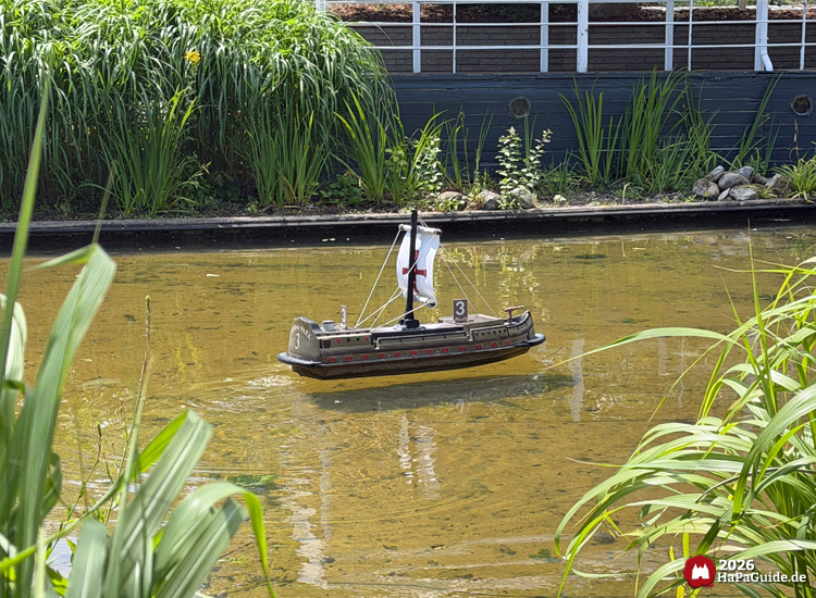 Modellschiff mit weißem Segel und rotem Kreuz fährt über den Teich im Hansa-Garten, umgeben von grünen Pflanzen und ruhigem Wasser, im Hintergrund dunkler Steg mit Geländer