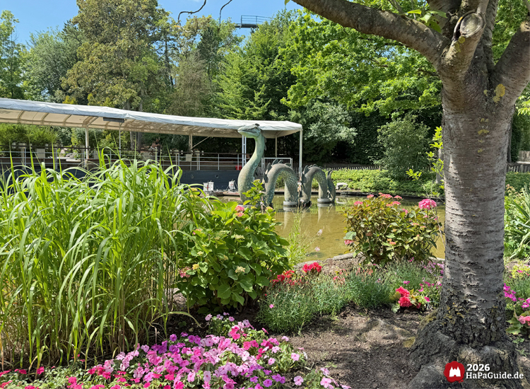 Blühende Gartenlandschaft am Teich der Fernlenkschiffe im Hansa-Park mit Seeungeheuer Nessie-Skulptur, pinken Blumen und hohen Gräsern, im Hintergrund überdachter Steg und Bäume