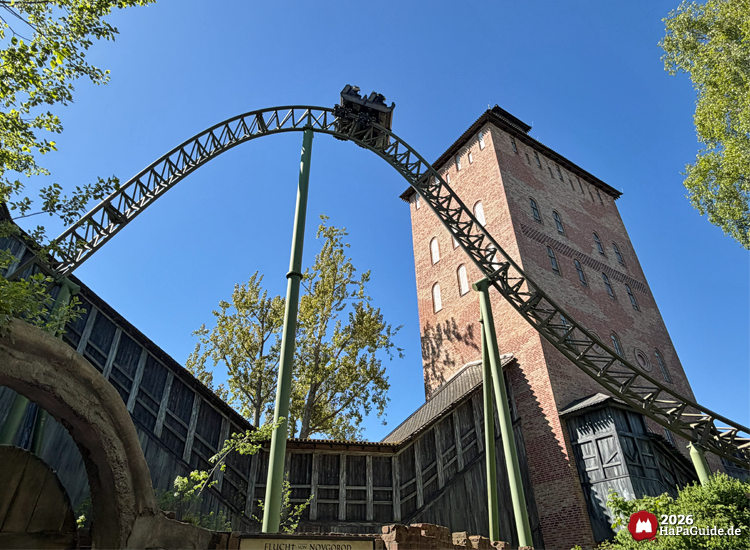 Achterbahn Flucht von Novgorod im Hansa-Park mit Zug auf dem Tophat vor dem Backsteinturm Saviour-Tower, umgeben von grünen Bäumen und blauem Himmel, zeigt den markanten Außenbereich der Anlage