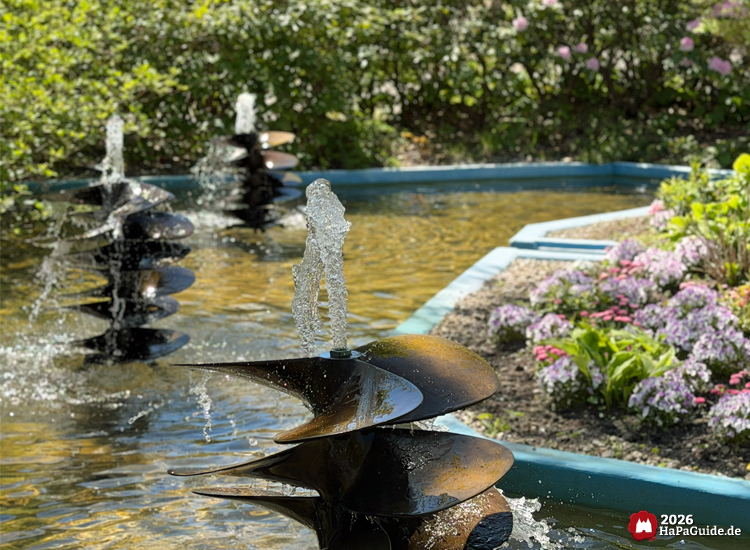 Dekorative Wasserfontänen mit spiralförmigen Schiffschrauben im Hansa-Park, umgeben von blühenden Blumenbeeten und grüner Vegetation bei sonnigem Frühlingswetter