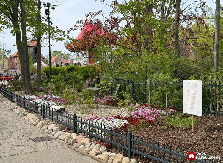 Blühender Hansa-Garten im Hansa-Park, umgeben von schwarzem Zaun und bunten Blumenbeeten, im Hintergrund das Kettenkarussell Hanse-Flieger mit rotem Dach