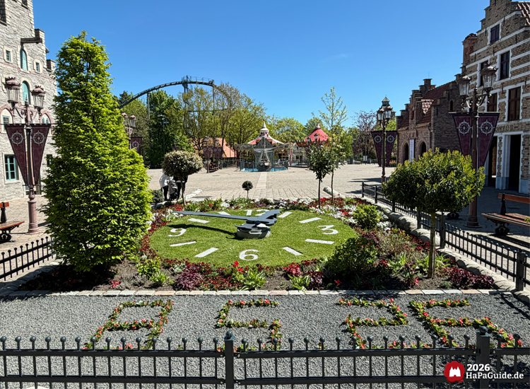 Blumenuhr im Themenbereich Hanse in Europa des Hansa-Parks mit gepflegtem Garten, grünen Sträuchern und Blick auf das Kettenkarussell Hanse-Flieger bei sonnigem Himmel
