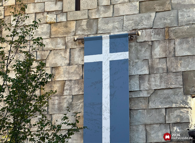 Detailaufnahme eines blauen Banners mit weißem Kreuz an der Steinwand des Palacio de Braga im Hansa-Park, daneben grüner Baum und strukturierte Steinmauer