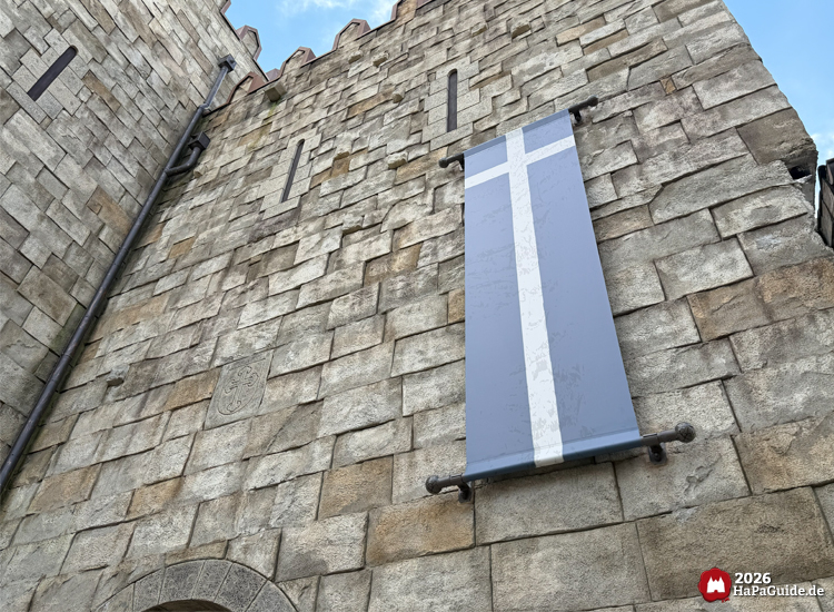 Detailaufnahme der Steinmauer des Palácio de Braga im Hansa-Park mit blauem Banner und weißem Kreuz, mittelalterliche Zinnen und Steinrelief unter blauem Himmel