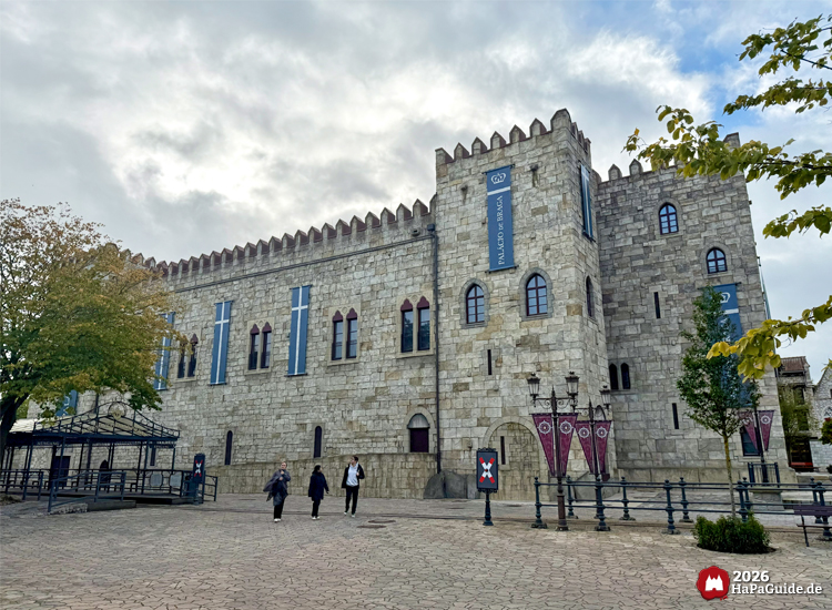 Seitenansicht des Palácio de Braga im Hansa-Park mit Zinnen, blauen Bannern und roten Fensterrahmen, Besuchende spazieren über gepflasterten Platz unter bewölktem Himmel