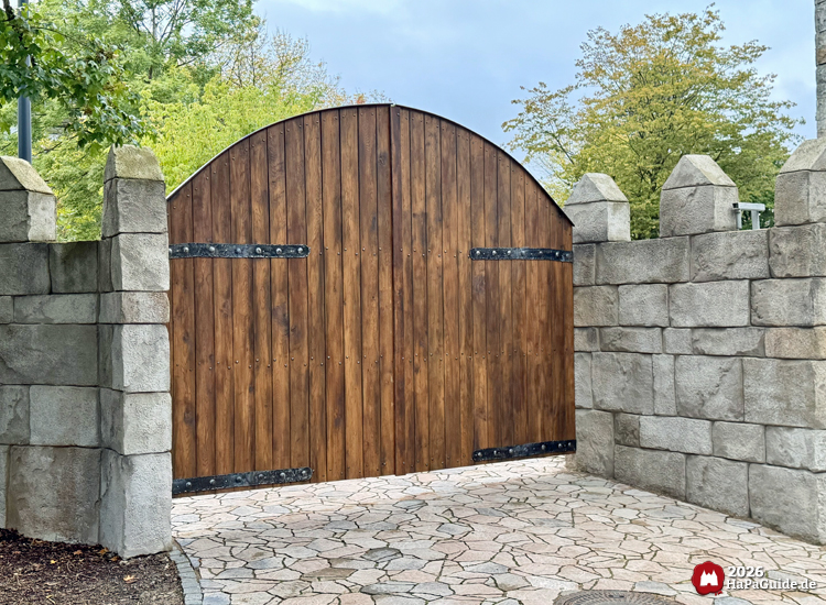 Großes hölzernes Tor mit schwarzen Metallbeschlägen hinter dem Palácio de Braga im Hansa-Park, eingebettet in graue Steinmauer mit Zinnen und umgeben von Bäumen im Herbstlaub