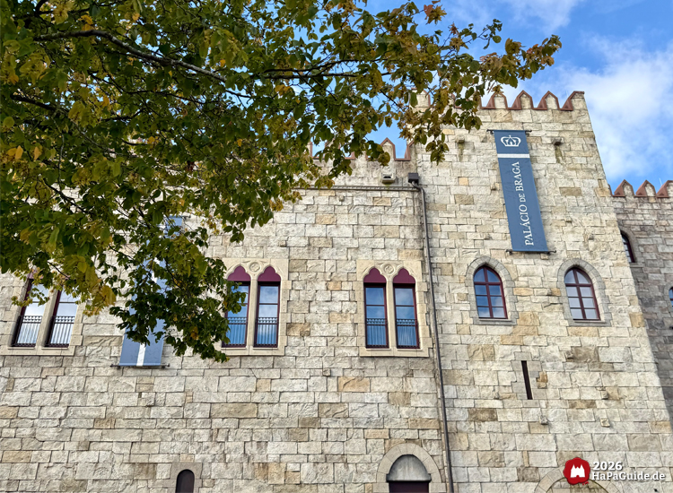Palácio de Braga im Hansa-Park mit roten Fensterrahmen und blauer Banneraufschrift, teilweise verdeckt durch grünen Baum im Vordergrund unter blauem Himmel