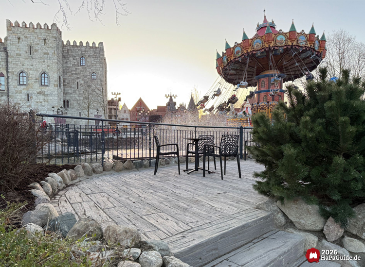 Sitzbereich auf Holzterrasse vor Zaun mit Blick auf schwingendes Kettenkarussell Hanse-Flieger und Burgkulisse Palacio de Braga im Hansa Park