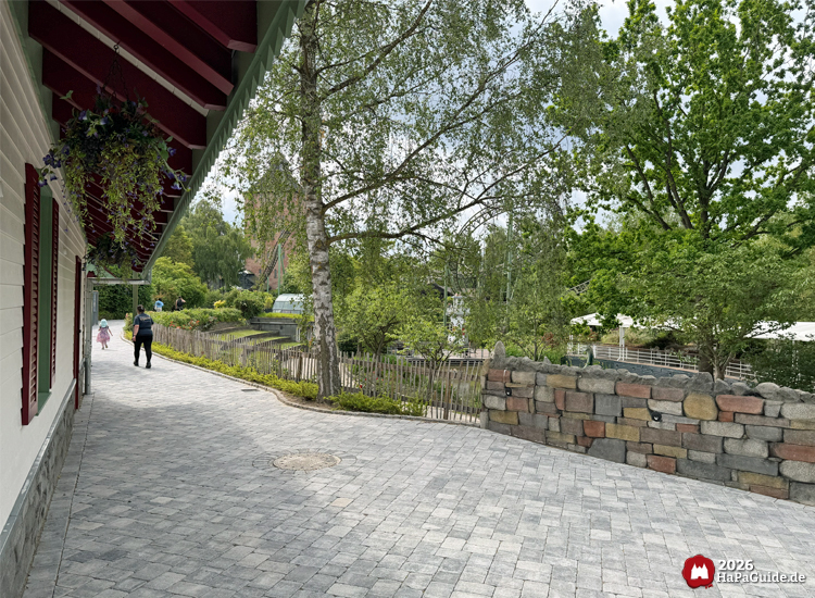 Gepflasterter Weg im Hansa-Garten mit Blick auf Bäume, Steinmauer und Teile der Achterbahn Flucht von Novgorod, Besuchende spazieren entlang des Gebäudes mit roten Fensterläden