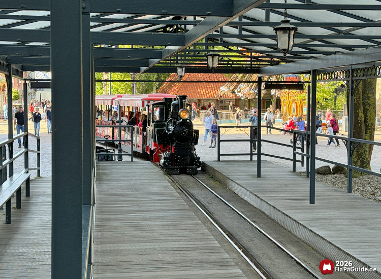 Dunkelgrüne und rote Dampflok des Hansa-Park Express fährt in die überdachte Station mit Holzplattform, Fahrgäste steigen ein, im Hintergrund Menschen am Carrousel Baltique