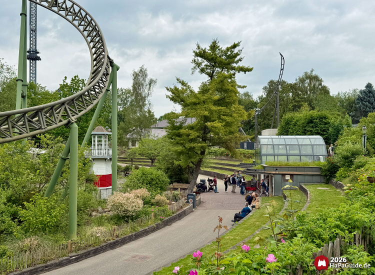 Grüne Schienen der Achterbahn Flucht von Novgorod im Hansa-Garten führen durch eine Kurve über den Weg, Besuchende spazieren entlang des Lachtempels und der bepflanzten Anlage