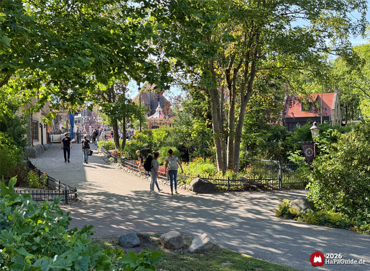 Spazierweg im Hansa-Garten mit Besuchenden zwischen grünen Bäumen und Blumenbeeten, im Hintergrund Gebäude mit roten Dächern