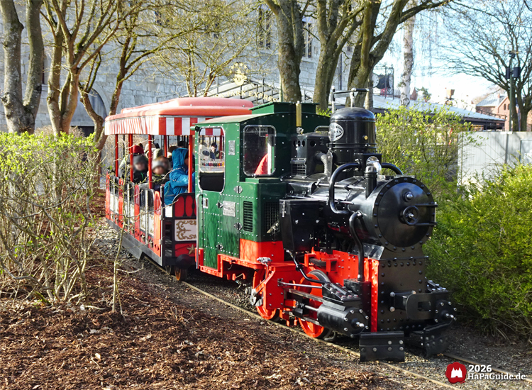 Dampflok Hansa-Park Express mit offenem Personenwagen fährt aus dem Hauptbahnhof Hansa-Garten durch einen begrünten Abschnitt mit Bäumen und Büschen.
