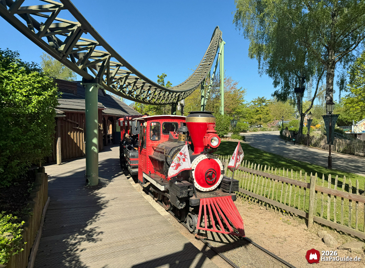 Rote Dampflokomotive der Parkbahn Hansa-Park Express im Hansa-Park mit schwarzen Details und Flaggen mit Hansa-Park Wappen, fährt unter der Achterbahn Flucht von Novgorod entlang des Bahnhofs im Peterhof bei sonnigem Wetter