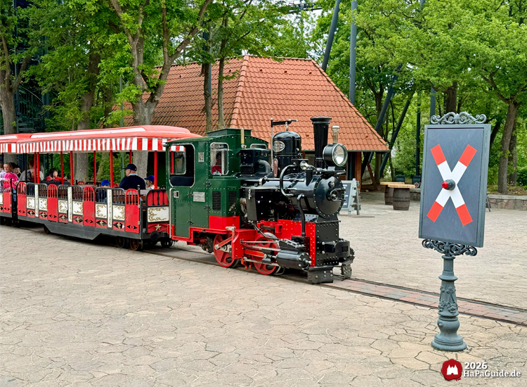 Grün-schwarze Dampflokomotive der Parkeisenbahn Hansa-Park Express im Hansa-Park mit roten Akzenten und rot-weiß gestreiften Waggons, überquert einen Bahnübergang der Hanse in Europa mit X-Schild auf gepflastertem Platz