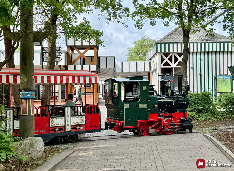 Grün-rote Dampflokomotive des Hansa-Park Express im Hansa-Park fährt durch den seitlichen Bereich des Bezaubernden Britanniens mit Bäumen und gestreiften Gebäuden, begleitet von rot-weiß gestreiften Waggons