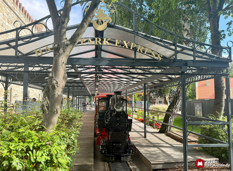 Station des Hansa-Park Express mit schwarzer Metallüberdachung und goldener Beschriftung, kleine Dampflok steht auf den Gleisen zwischen Bäumen und Blumenbeeten