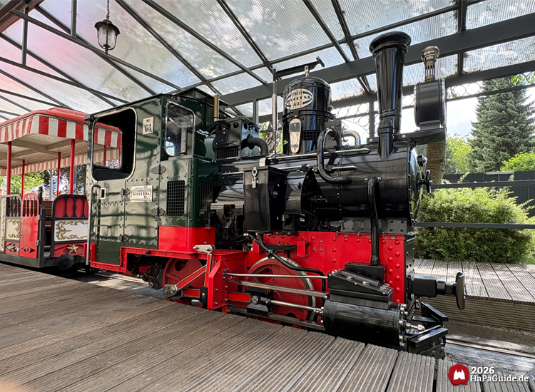 Schwarz-rote Dampflok des Hansa-Park Express unter Glasdach, grüne Führerkabine mit Messingschildern und angehängtem rot-weißem Wagen im Hintergrund