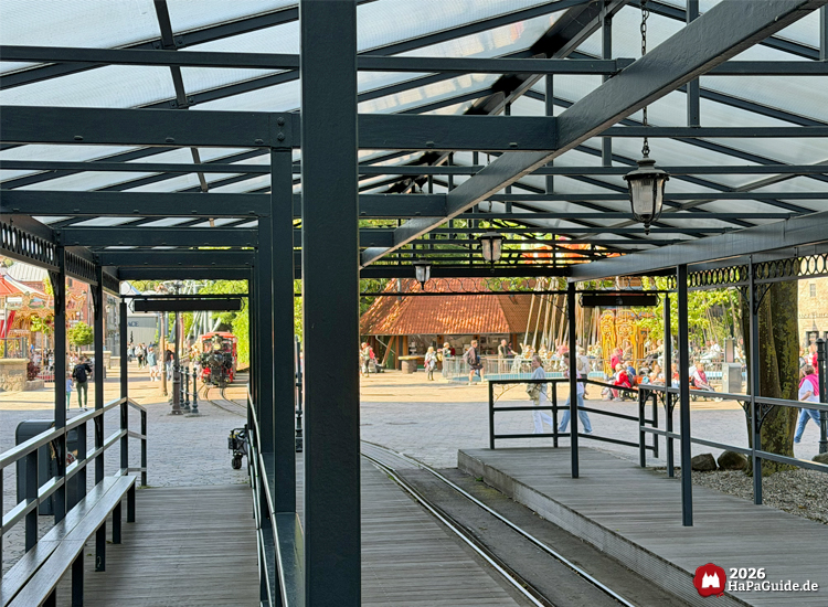 Blick von der überdachten Station des Hansa-Park Express auf die Gleise und einen Platz mit Besuchenden in der Hanse in Europa, im Hintergrund die Lokomotive und der Kiosk am Hanse-Flieger