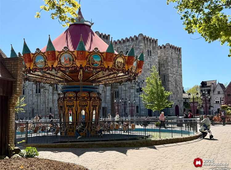 Kettenkarussell Hanse-Flieger im Hansa-Park mit rotem Dach und goldenen Verzierungen vor der Kulisse des mittelalterlichen Palacio de Bragas und gepflastertem Platz, Besuchende genießen die sonnige Atmosphäre