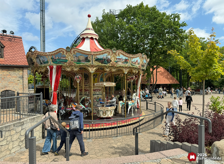 Hanse-Karussell im Hansa-Park mit roten und weißen Streifen am Dach, goldenen Verzierungen und bemalten Tafeln, Besuchende spazieren über den Platz mit Bäumen und Sitzbänken