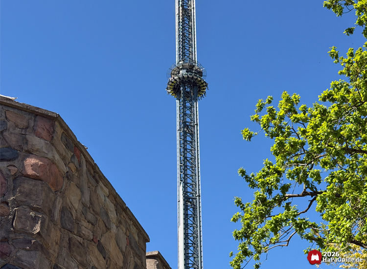 Hoher Freifallturm Highlander im Hansa-Park mit Fahrgästen in luftiger Höhe, daneben Steinmauer und grüner Baum unter blauem Himmel