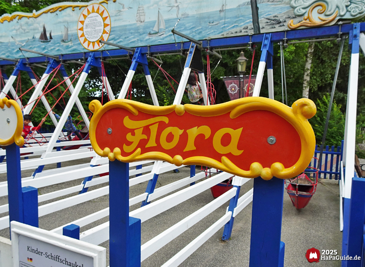 Kinder-Schiffschaukel Flora im Hansa-Park mit rot-gelbem Namensschild, blauen und weißen Holzstreben und maritimer Bemalung mit Segelschiffen im Hintergrund