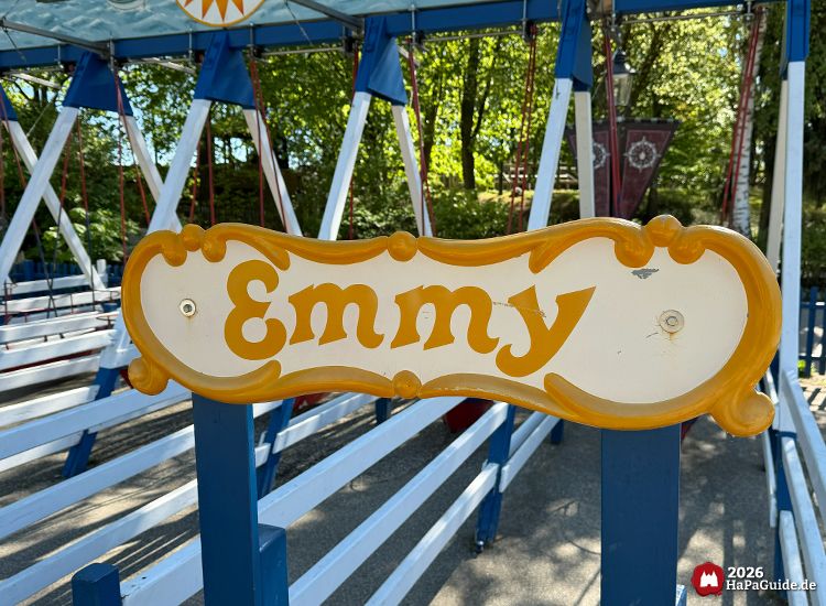 Kinder-Schiffschaukel Emmy im Hansa-Park mit cremefarbenem Schild und gelber Verzierung, blauen Holzstreben und roten Sitzbooten unter grünen Bäumen
