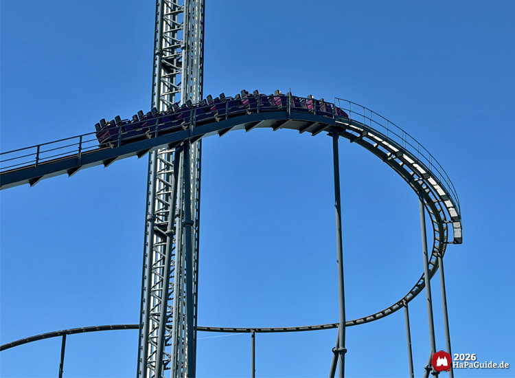 Lila Achterbahnzug Nessies im Hansa-Park erklimmt den Lift vor dem Stahlturm des Highlanders bei klarem blauem Himmel