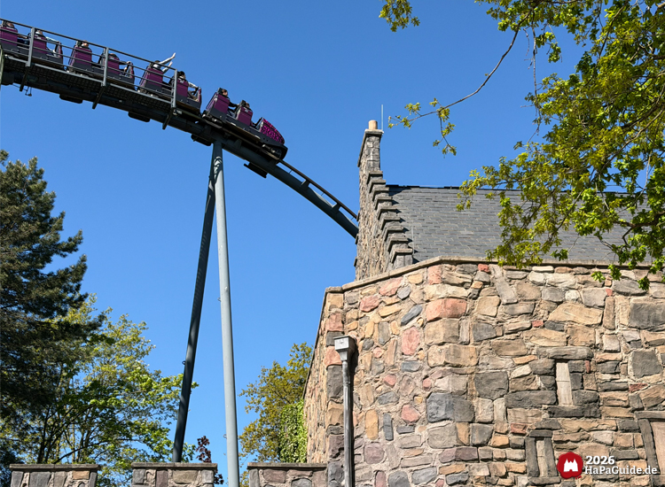 Lila Achterbahnzug von Nessie im Hansa-Park fährt mit Airtime neben dem Schieferdach des Eilean Donan Castles abwärts unter blauem Himmel