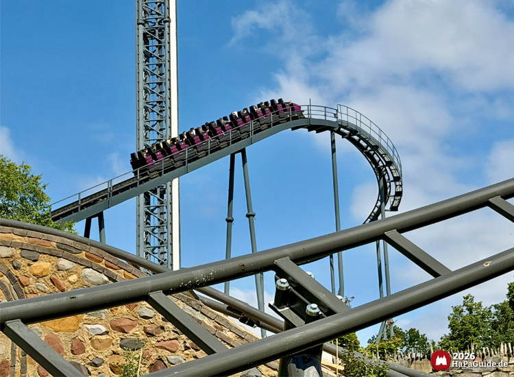 Zug der Loopingachterbahn Nessie im Hansa-Park fährt den steilen Lifthill hinauf, schwarze Stahlkonstruktion mit grauen Stützen und Steinmauer im Vordergrund unter blauem Himmel