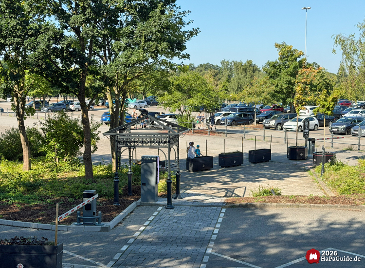 Eingangsbereich des Hansa-Park Parkplatzes mit Automat unter dekorativem Metalltor, Pflanzkübeln und geordneten Parkreihen im Hintergrund