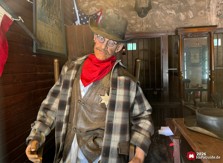 Sheriff-Animatronic im Westernstil im Hansa-Park mit Cowboyhut, rotem Halstuch und Lederweste, sitzt in einem rustikalen Büro mit Holzinterieur und amerikanischer Flagge im Hintergrund