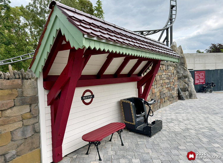 Thematisierte Bushaltestelle im Hansa-Park mit rotem Dach, grünen Zierleisten und weißer Wand mit 'Bus Stop'-Schild, daneben eine rote Bank und ein Test-Sitz für die Achterbahn Cornwall Coaster