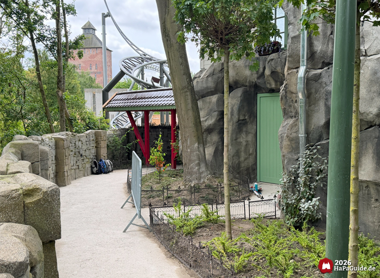 Ausgangsweg im Hansa-Park entlang künstlicher Felsen mit grüner Tür im Hintergrund, dahinter die Achterbahn Cornwall Coaster und der Saviour-Tower der Flucht von Novgorod unter bewölktem Himmel