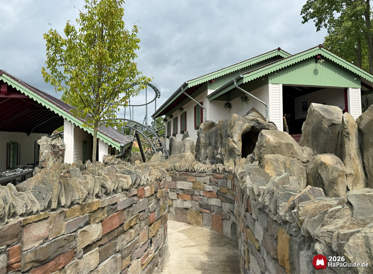 Blick auf die Gebäude der Achterbahn Cornwall Coaster im Hansa-Park mit weißen Fassaden, grünen Dachkanten und roten Fensterläden, umgeben von Felsen und einem gepflasterten Weg des Wartebereichs