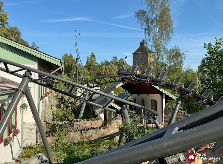 Zug der Cornwall Coaster Achterbahn im Hansa-Park fährt durch geschwungene schwarze Schienen zwischen Gebäuden mit grünem Dach und rotem Rahmen, im Hintergrund der Turm der Flucht von Novgorod