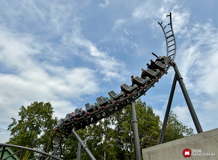 Zug des Cornwall Coaster im Hansa-Park fährt auf den Spike welcher 2027 zum Roche Rock weitergestaltet wird, Fahrgäste genießen die Fahrt unter blauem Himmel mit Wolken und grünen Bäumen