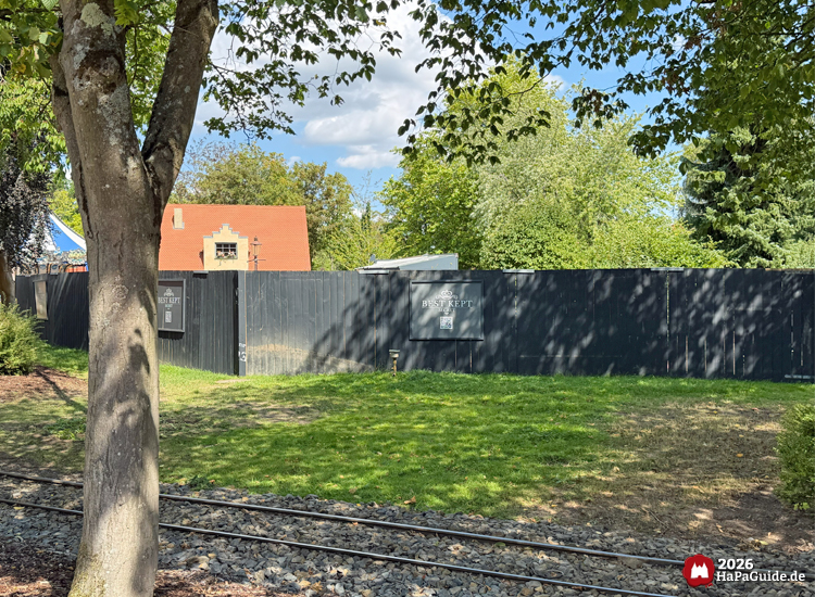 Schwarzer Bauzaun mit Beschriftung 'Best Kept Secret' im Hansa-Park, dahinter Bäume und Gebäude mit rotem Dach, im Vordergrund Schienen der Parkbahn und grüne Wiese, Bauplatz Neuheit 2027