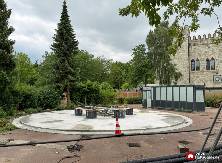 Runde Betonplattform mit schwarzer Glaskonstruktion des ehemaligen Holsteinturms im Bauabschnitt Hansa-Park Neuheit BKS2 2027, im Hintergrund Fassade des Palacio de Braga mit Zinnen und roten Fenstern, umgeben von Bäumen