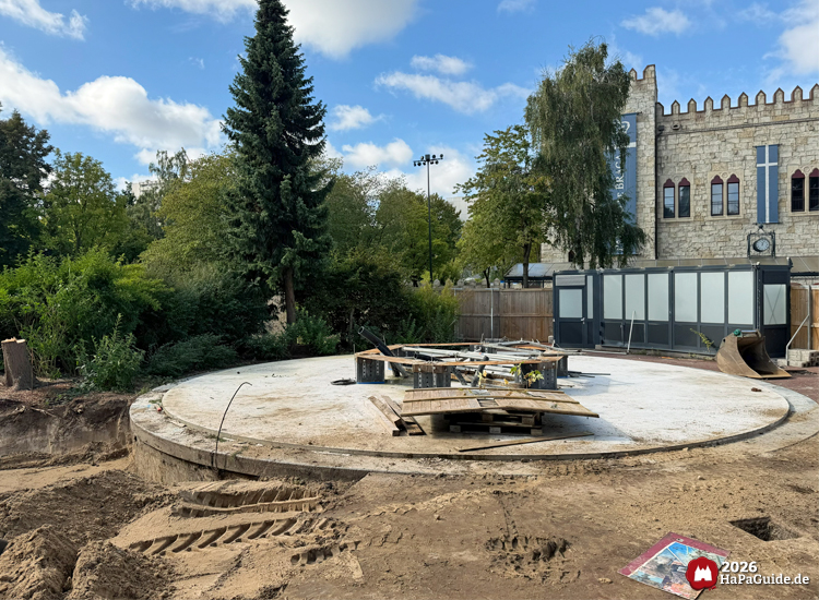 Baustelle des Buddenbrook-Turms im Hansa-Park mit rundem Betonfundament und Baumaterialien, im Hintergrund das steinerne Gebäude des Palácio de Braga mit roten Fenstern und blauen Bannern