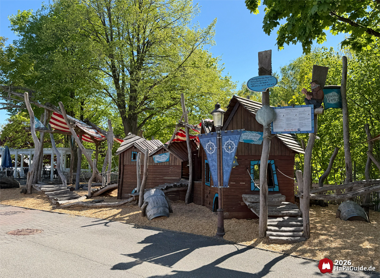 Niedrigseilgarten im Hansa-Park mit rustikalen Holzhäusern, roten und weißen Segeltüchern und blauen Schildern, umgeben von grünen Bäumen bei Sonnenschein