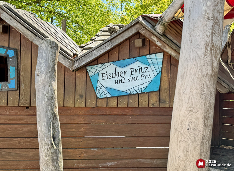 Holzschild mit der Aufschrift 'Fischer Fritz und sine Fru' an der Fassade eines braunen Holzhauses am Hansa-Park Niedrigseilgarten, gestaltet mit blauer Fischernetz-Optik und maritimen Details