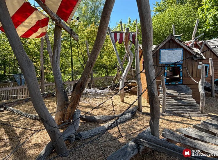 Innenbereich des Spielplatzes Niedrigseilgarten im Hansa-Park mit Holzstegen, Kletterseilen und roten Segeltüchern, umgeben von Bäumen und Holzspänen